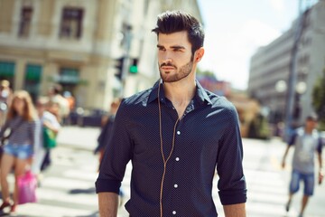 Fototapeta premium Handsome young man walking in the city, looking away, listening to music through earbuds.