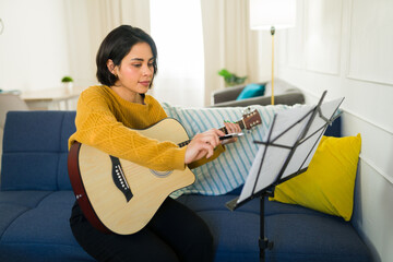 Focused young woman practicing guitar chords at home, using a music stand to follow notes and...