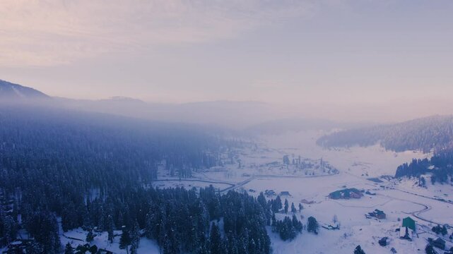 aerial drone shot flying forward over a fog, haze, cloud covered gulmarg valley with fresh snowfall, coniferous chinar walnut trees and the sunlight coming from behind himalaya mountains in Kashmir