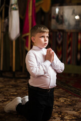 A young boy, dressed in a white shirt and dark pants, kneels in prayer with his hands clasped. The...