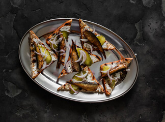 Delicious appetizer, tapas, snack - sandwiches with rye bread, canned sardines, gherkins and cream cheese on a dark background, top view