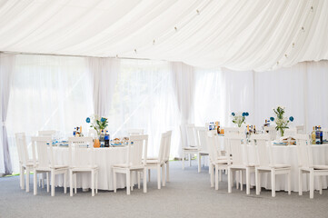A beautifully decorated event tent with white tables and chairs.  Blue flowers adorn each table,...