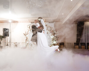A bride and groom share their first dance, enveloped in a romantic fog effect. The wedding reception venue is elegantly decorated. A magical, unforgettable moment.
