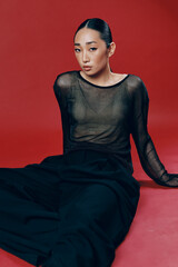 Fashion asian woman in black pants and sheer top sitting on red surface with legs crossed and hands...