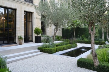 Obraz premium Beautiful garden with olive trees, serene water feature, and well-manicured hedges next to a stylish modern home in the early evening light