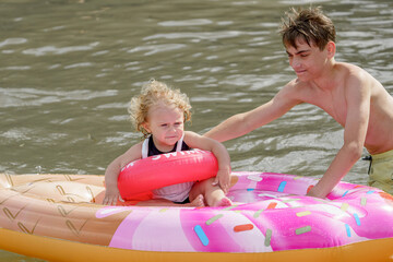Fototapeta premium A little girl with an inflatable ring swims with her brother on an inflatable mattress and floats in the water at sea.