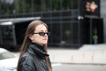 Fototapeta premium A woman with long hair wearing dark sunglasses stands confidently on a city street. She is dressed in a black jacket, with a sleek, contemporary building in the background
