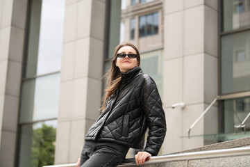Obraz premium A young woman in a black quilted jacket and sunglasses relaxes against a railing. The backdrop features a contemporary building and a clear sky, capturing a casual urban lifestyle