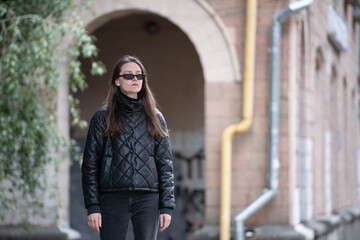 A young woman dressed in a black puffer jacket and sunglasses stands confidently by a brick...