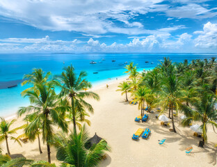 Aerial view of white sandy beach with palm trees, sunbeds, umbrellas, yachts, boats, blue ocean, sky with clouds at sunset. Summer in Kendwa, Zanzibar island. Tropical landscape. Clear sea. Top view