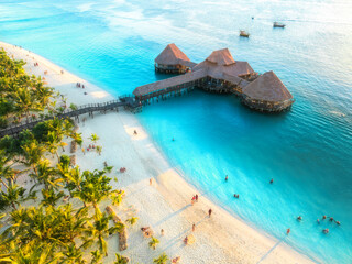 Aerial view of beautiful bungalow in Indian ocean at sunset in summer. Restaurant on the sea. Top view of wooden hotel, palm trees, azure water, white sandy beach. Luxury resort in Kendwa, Zanzibar