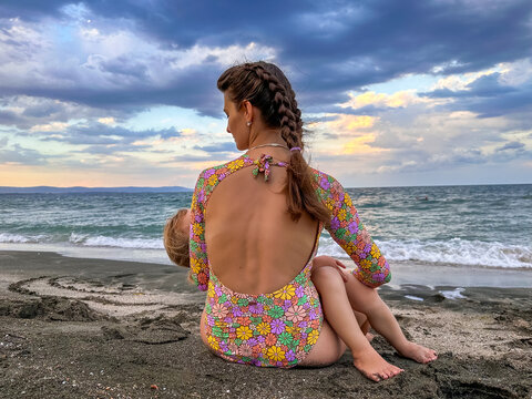 Mom is sitting on the seashore in a swimsuit and holding her sleeping baby. Bright swimsuit and waves on the sea. Aheloy, Bulgaria