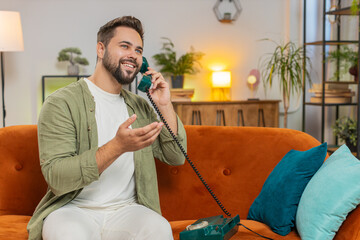 Smiling young man making wired telephone call conversation with friends sitting on couch at home....