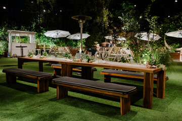 Outdoor dining setting with long wooden tables and benches, decorated with greenery under patio umbrellas at night.