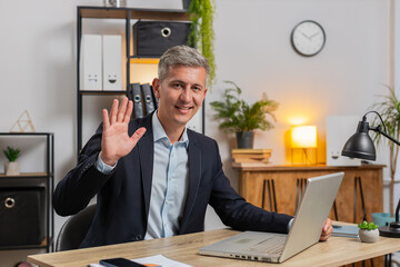 Hello. Happy young businessman employee waves hand hi gesture greeting welcomes someone, online webinar, remote distance conversation. Freelancer guy in suit sitting at desk with laptop in home office © Andrii Iemelianenko
