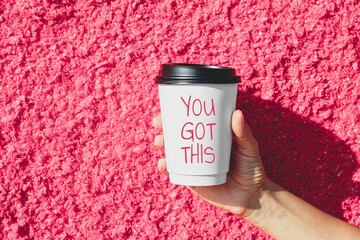 A woman hand holding a white takeaway coffee cup with motivational text "You Got This" against a vibrant pink textured background, conveying positivity and energy
