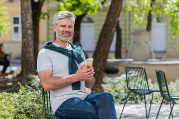 Fototapeta premium Happy smiling mature man using smartphone typing text messages tired looking up thinking and browsing internet social media web app working chatting online outdoor. Joyful guy in urban city street