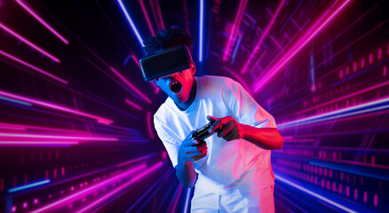 Obraz premium Teenager holding joystick and wearing VR goggles while enjoy playing games at neon abstract background. African man enjoy controlling controller and using augmented reality technology. Ingenuity.