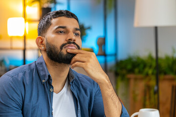 Bearded young Indian male freelancer in smart casual thinking while sitting with laptop at home office. Thoughtful Arabian business man thinking about an important creative project work decision.