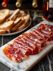 Sliced cured meat is neatly arranged on a marble board, next to a plate of bread, with wine bottles in the background. Concept of gourmet dining setup. For food-related content