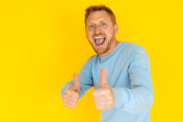 Excited redhead man smiling widely and giving a thumbs up against a vibrant yellow background, radiating positivity and joy