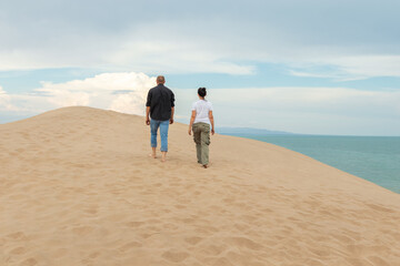 Two People Walking on Sandy Dune Ridge Overlooking Ocean Horizon Under Cloudy Sky at Coastal Landscape