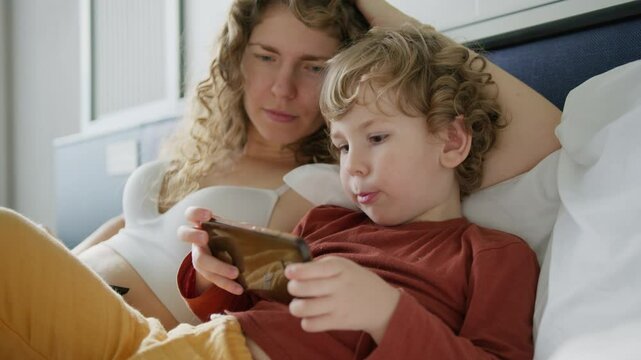 Loving mother and her son are enjoying a cozy moment together while engaging with a device on their bed. Sharing a Beautiful and Heartwarming Moment Together