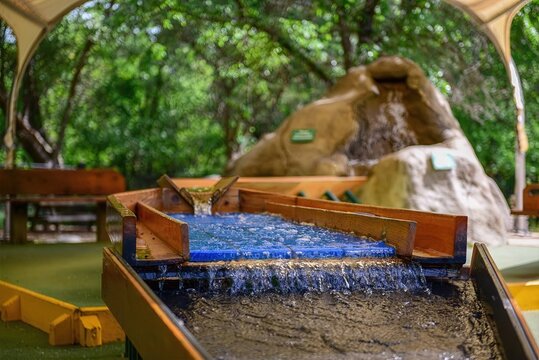 Mining sluice boxes in a shady grove, water rushing over rocks, re-creating gold panning experience in a lush setting.

