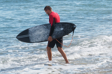A skilled surfer steps in of the water, holding a black surfboard, as waves lap at his feet.