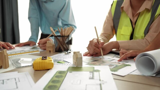 Smart architect and safety engineer discussion about building blueprint on table with architectural document, measurement tools and wooden block scatter around at modern office. Closeup. Delineation.