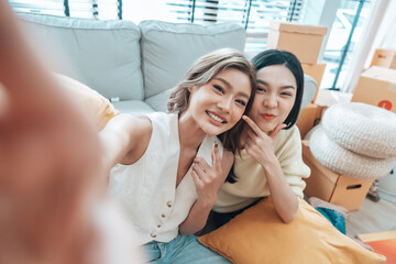 Two young Asian women smiling and happy while taking a selfie in their living room at home, looking at the camera