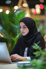 A young Muslim woman in a hijab is sitting at her laptop and working on digital marketing. The modern office background features green plants and flowers, blurred bokeh lights.