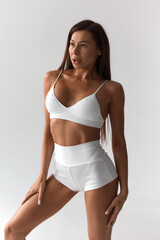 Obraz premium Tanned girl with athletic figure in white clothes warming up before training. Athletic girl posing in bright studio. Healthy lifestyle