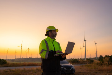 Engineer working on laptop in front of wind turbines for checking wind turbines of the field during beautiful sunset. Alternative energy concept.