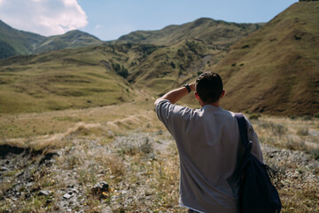 A man is a tourist in a gray suit and with a backpack against the background of green mountains in Georgia. Man is white, put his hand to his forehead and looks into the distance like a captain look