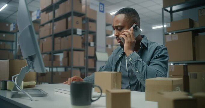 Sorting center: Male African American warehouse associate talks to customer by mobile phone, checks inventory on computer, prepares package for delivery. Storage with orders of online E-commerce store