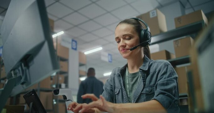 Female logistics manager in headset talks on call with customer, uses computer. Sorting center worker checking cardboard boxes for shipping to clients. Warehouse of postal service or E-commerce store.