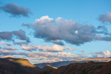 Mountain and hill tops illuminated by sunset light. Rocks silhouettes and gold sunrise colors. Vivid golden sunlight on hills under clouds of sunset tones in blue sky. Shadows of clouds on rocky ridge