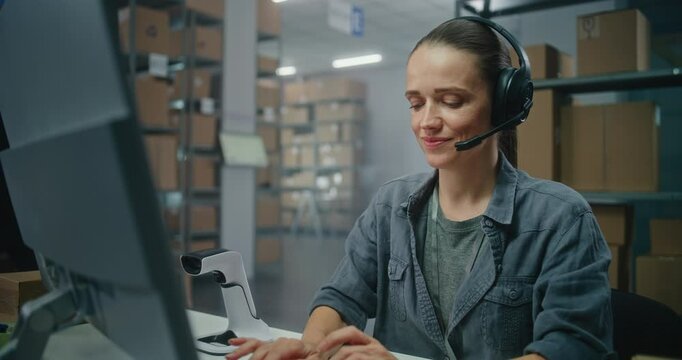 Warehouse of postal service or E-commerce store: Female logistics manager in headset talks on call with customer, uses computer. Sorting center worker checking cardboard boxes for shipping to clients.