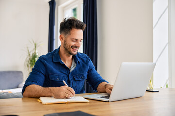 Adult man working at home using laptop taking notes in notepad