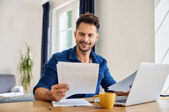 Happy man looking at document while working on laptop from home at living room