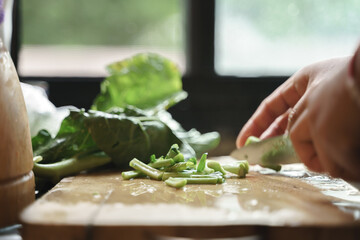 Close-up of hands chopping fresh green vegetables on a wooden cutting board with leafy greens in the background.