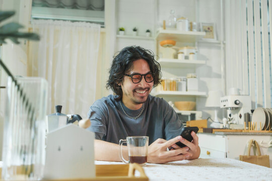 Asian man smiling while enjoying coffee and using a smartphone at the dining table, savoring a relaxed morning at home.