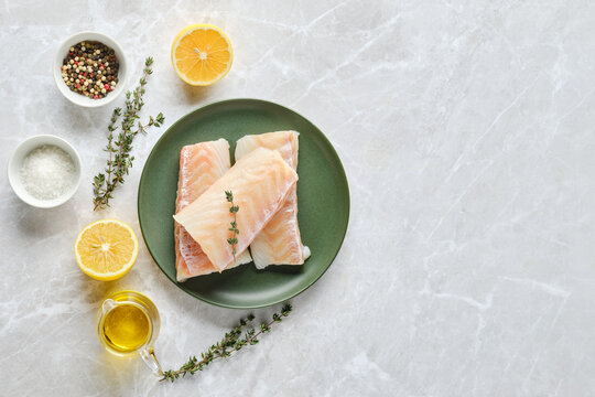 Overhead view of fresh cod fish fillets on a plate with herbs and citrus on a marble countertop