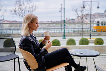 A woman enjoys a moment of relaxation while sipping coffee at an outdoor cafe, surrounded by a scenic urban view. Her leisure time captures a sense of tranquility and escape from the routine.