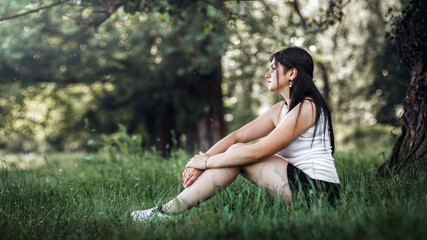 woman sitting on the grass in a park or forest. She is wearing a white T-shirt and black shorts. A woman sits with her arms wrapped around her knees and looks into the distance.