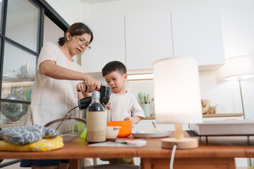 Asian mother teaching her young son how to use a hand mixer to make cookies in a bright modern kitchen with various baking tools and ingredients placed on the wooden table.