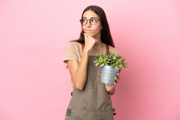 Young gardener girl holding a plant isolated on pink background having doubts and thinking