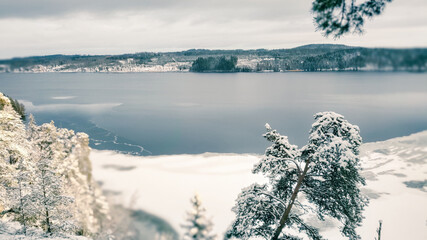 a winter landscape where a beautiful lake stretches across the scene. The center of the lake is still open water, reflecting the gray winter sky and the snow-covered trees that line its edges. On one 