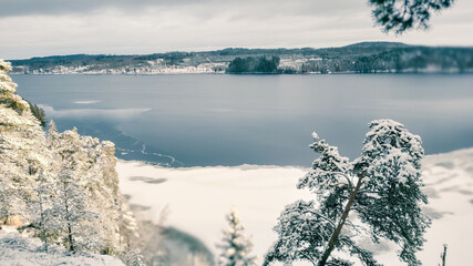 a winter landscape where a beautiful lake stretches across the scene. The center of the lake is still open water, reflecting the gray winter sky and the snow-covered trees that line its edges. On one 